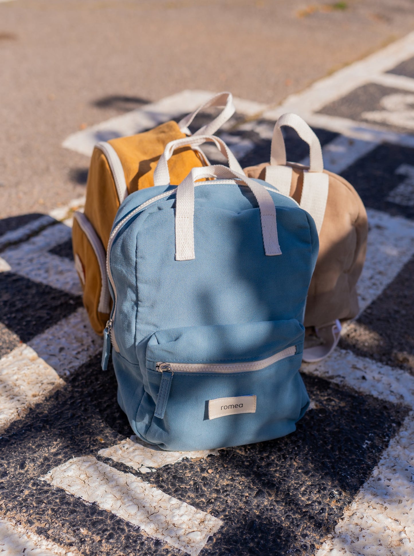 trois sacs à dos de couleur beige, moutarde et bleu posé dans une cour de récréation