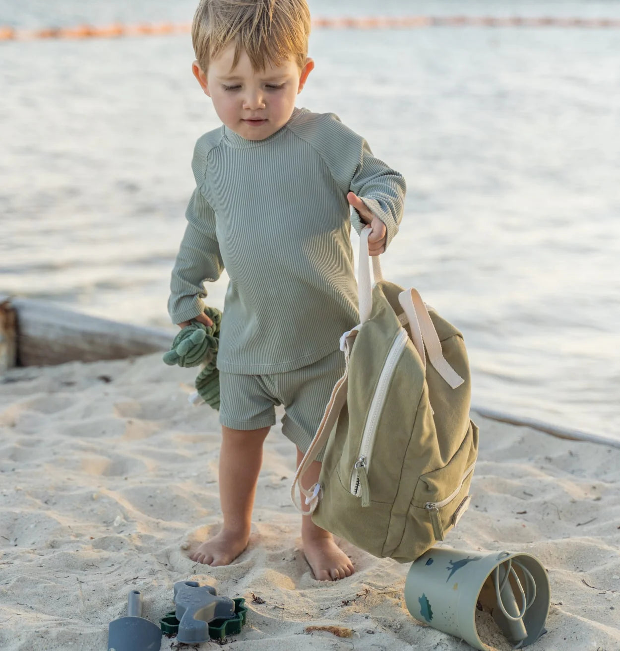 un petit garçon porte un sac à dos de couleur vert sur une plage 