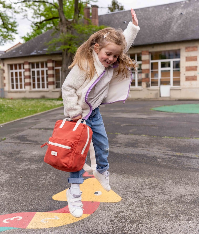 petite fille qui saute avec un sac à dos romea rouge brique 